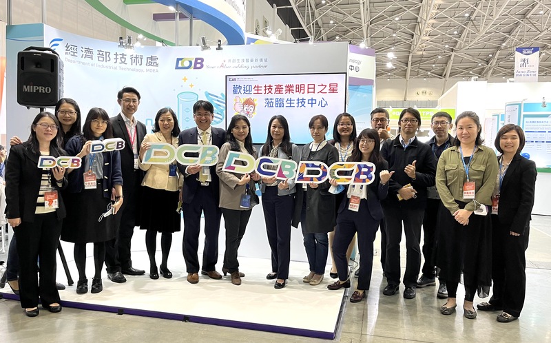 這張圖片顯示了一群人在展覽攤位前合照。他們手持著印有 "DCB" 字樣的標牌，背景看起來是一個科技展覽的攤位。
