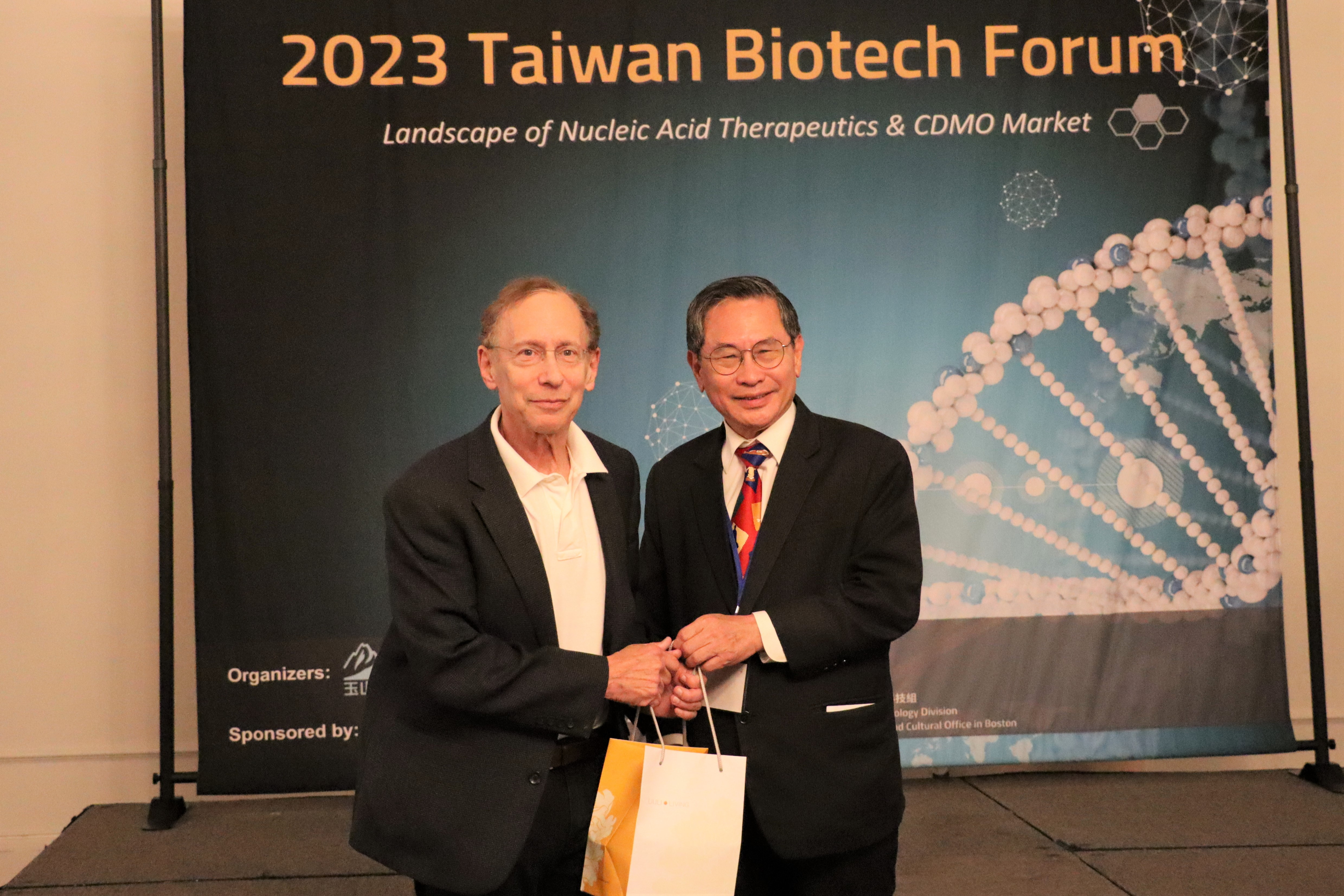 圖片中有兩位身穿西裝的男性站在一起，背景是一個關於2023年台灣生技論壇的海報，海報上寫著「2023 Taiwan Biotech Forum」以及「Landscape of Nucleic Acid Therapeutics & CDMO Market」。