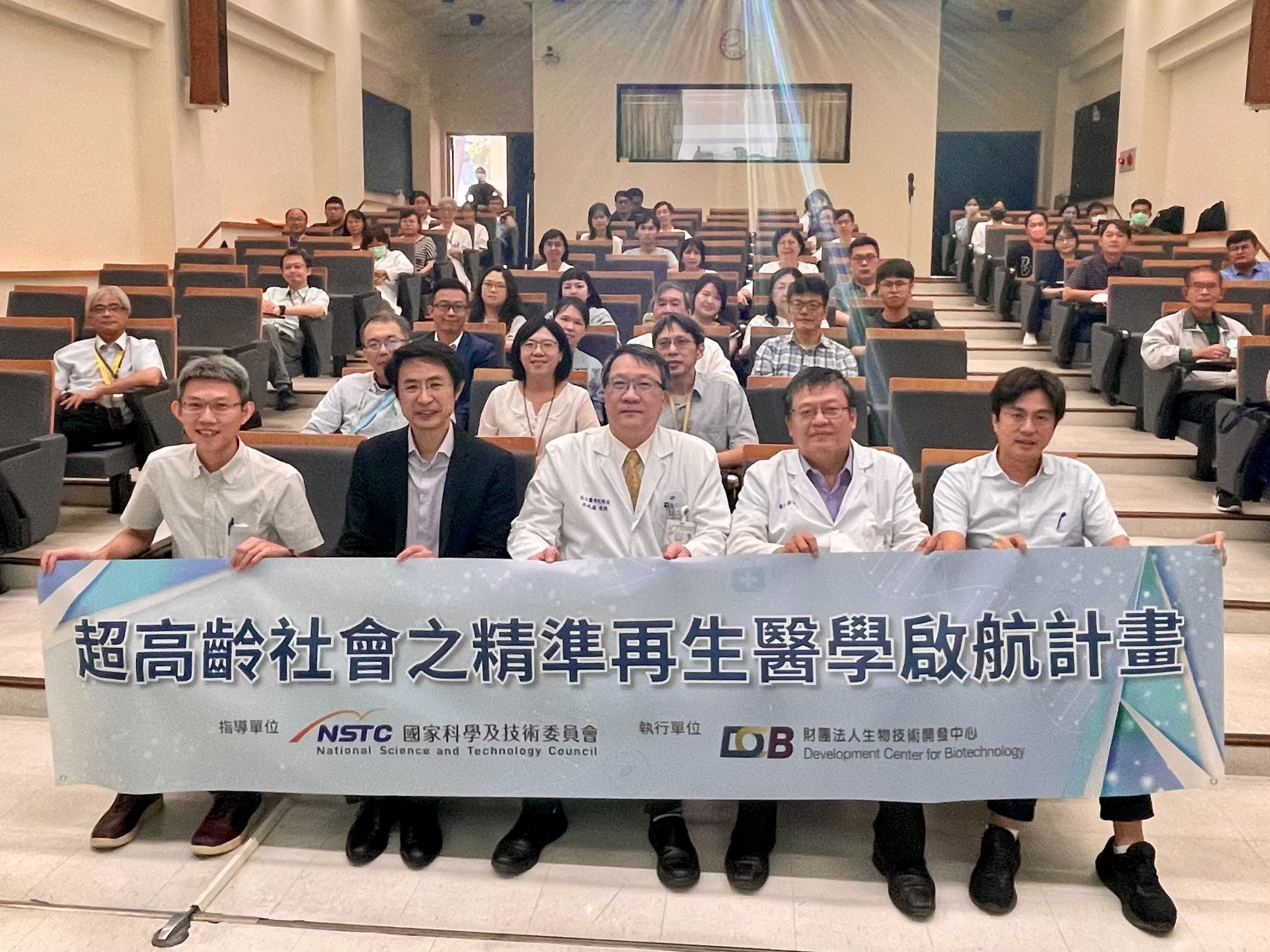 這張圖片顯示一群人在一個教室裡，他們坐在階梯教室的座位上，前排的人拿著一個橫幅，上面寫著“超高齡社會之精準再生醫學啟航計畫”。