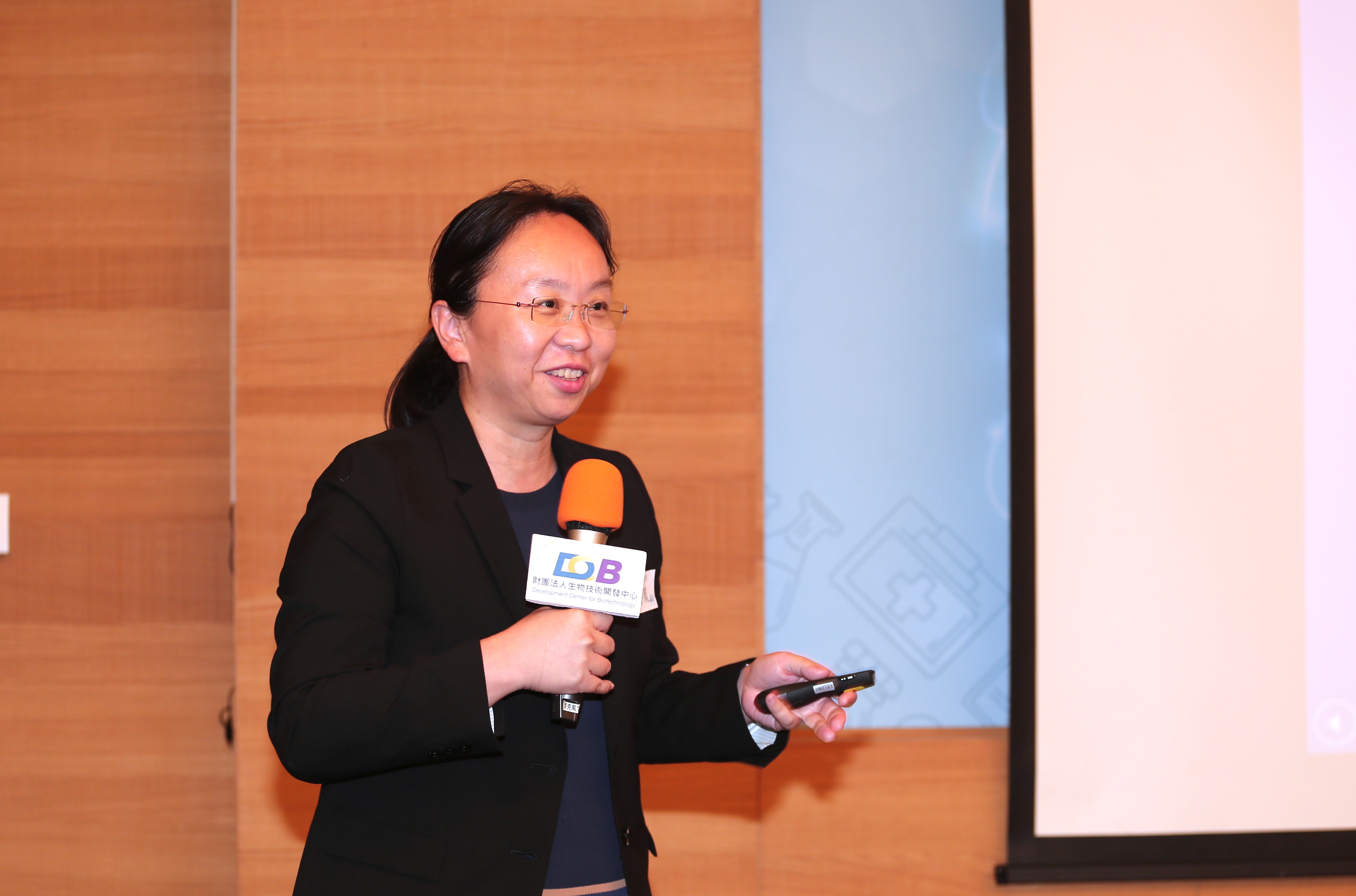 這張圖片展示了一位穿著黑色西裝的女士，她正在進行演講。她手持一個橙色麥克風，並且拿著一個遙控器。背景是一個木質牆面和一個投影屏幕。