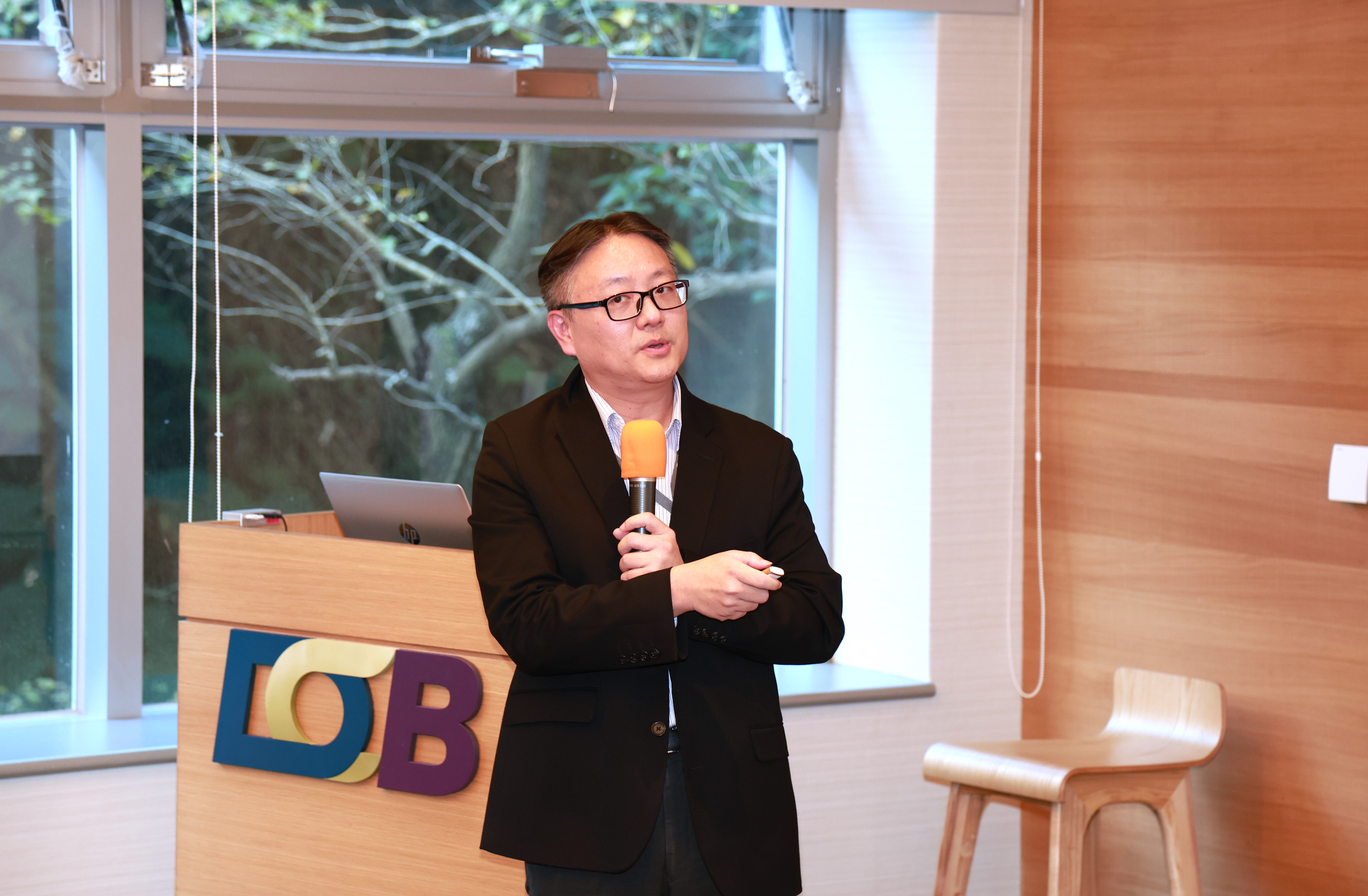 這張圖片顯示一位身穿黑色西裝的男士正在進行演講。他手持橙色麥克風，並且站在一個講台旁邊。講台上放置了一台筆記型電腦，背景是一扇大窗戶，窗外可以看到一些樹木的景色。