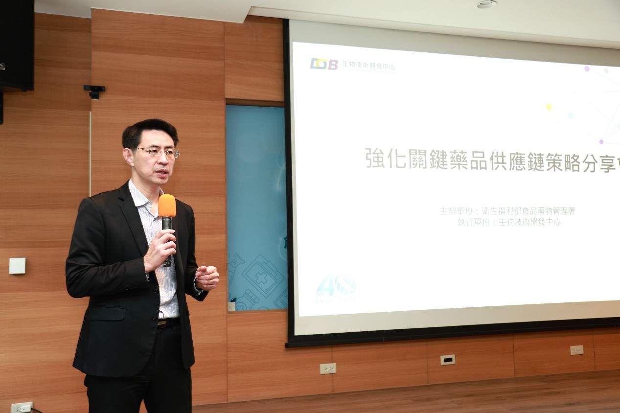 這張圖片中，一位穿著黑色西裝的男子正拿著麥克風發表演講。他站在一個木質背景前，旁邊有一個投影屏幕。