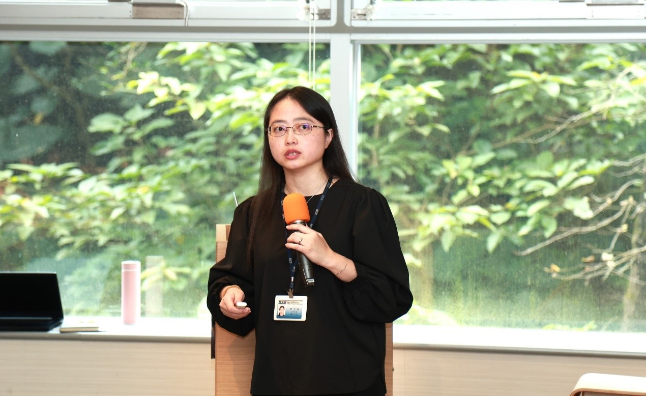 這張圖片顯示一位女性正在進行演講，她手持橙色麥克風，穿著黑色上衣，佩戴識別證。背景是一扇大窗戶，窗外有茂密的綠色植物。