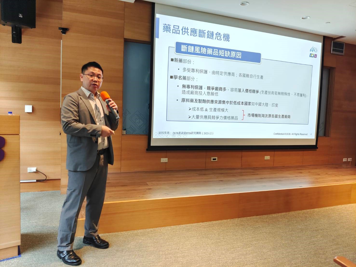 這張圖片顯示一位穿著西裝的男子正在進行演講，他手持麥克風，站在一個投影屏幕前。屏幕上顯示的內容與藥品供應鏈斷鏈危機有關，並列出了斷鏈風險藥品短缺原因。