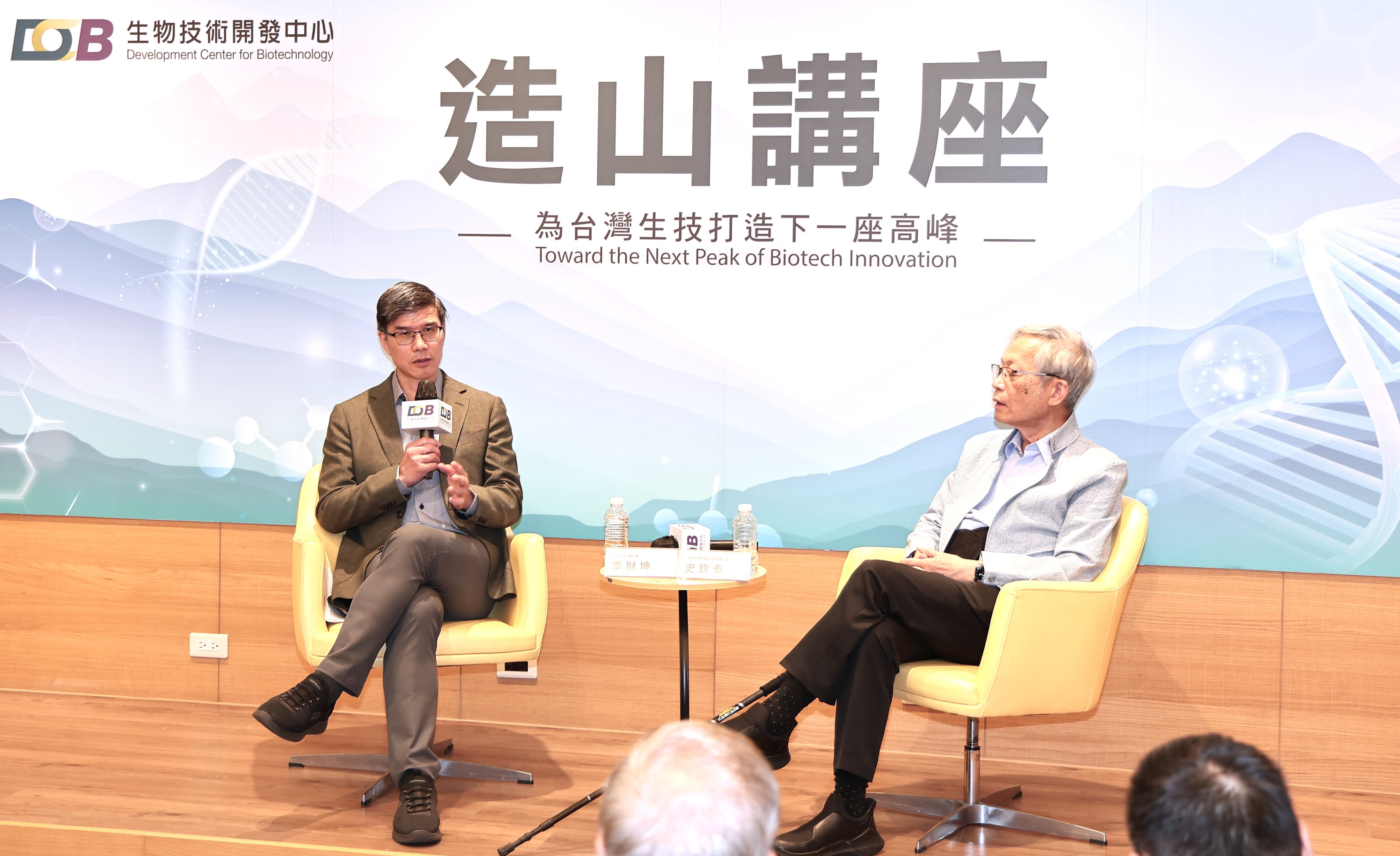 圖片呈現一場生物科技主題的公開論壇或座談會，舞台背景印有「生物技術開創新高峰」及「邁向下一個生技高峰」等標語，並搭配英文口號。中央一名穿著西裝的男士手持麥克風發言，兩側可見其他與談人員及桌上的瓶裝水。背景採用藍白色科技感圖案，現場前方坐著觀眾，整體氛圍正式且具學術交流與產業發展特色。