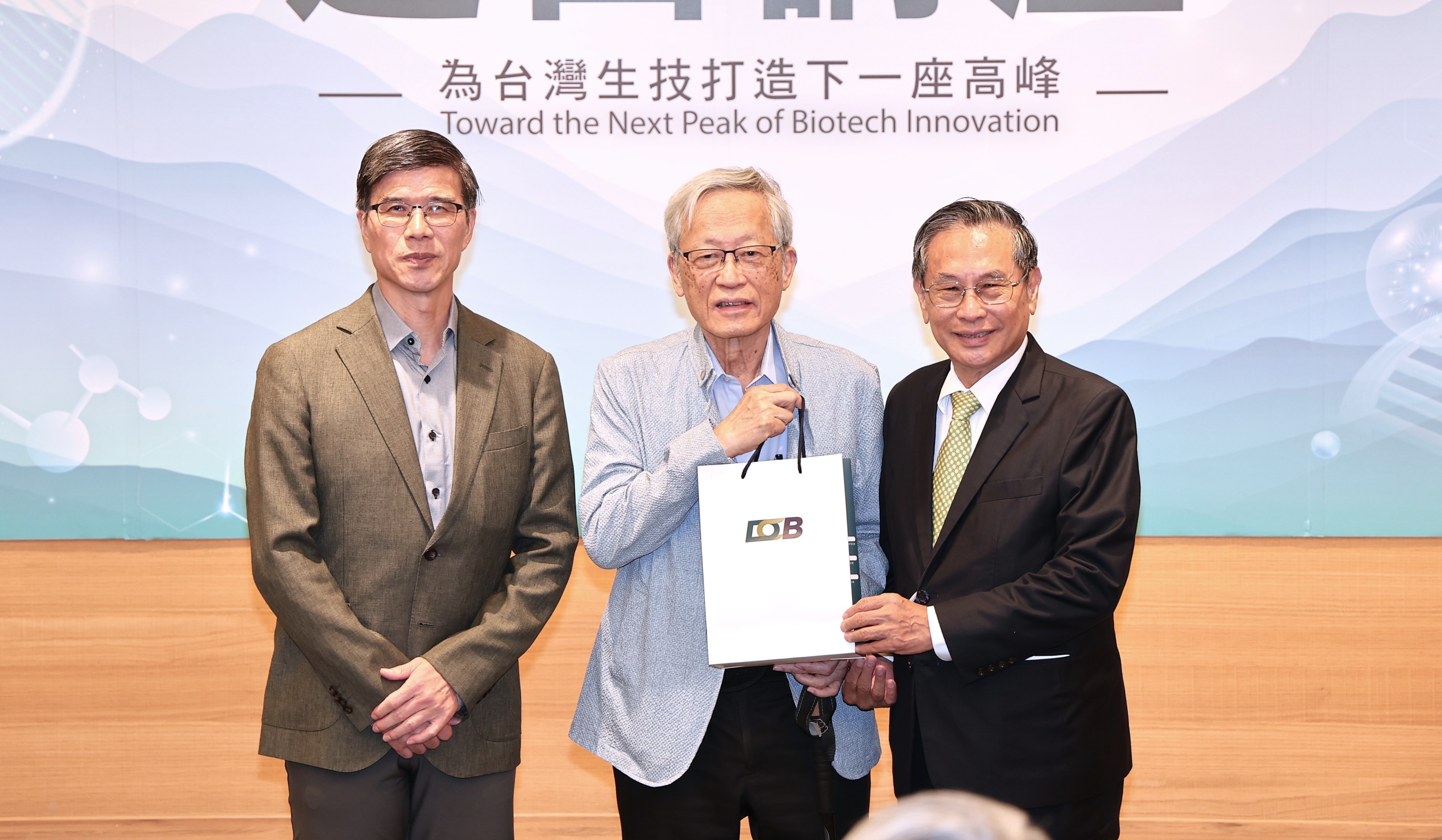 三位年長男性在活動或論壇現場合影，皆穿著正式西裝，其中兩人戴著眼鏡，表情自然親切。背景是一面大型投影或布幕，上方印有中英文標題，內容似乎與生物科技、創新發展或下一個突破高峰有關。現場燈光明亮，地面與桌面呈現溫暖木質色調，整體氣氛專業而隆重。右側人士手持一張大型證書或海報，畫面展現團隊合作與學術交流的意義。