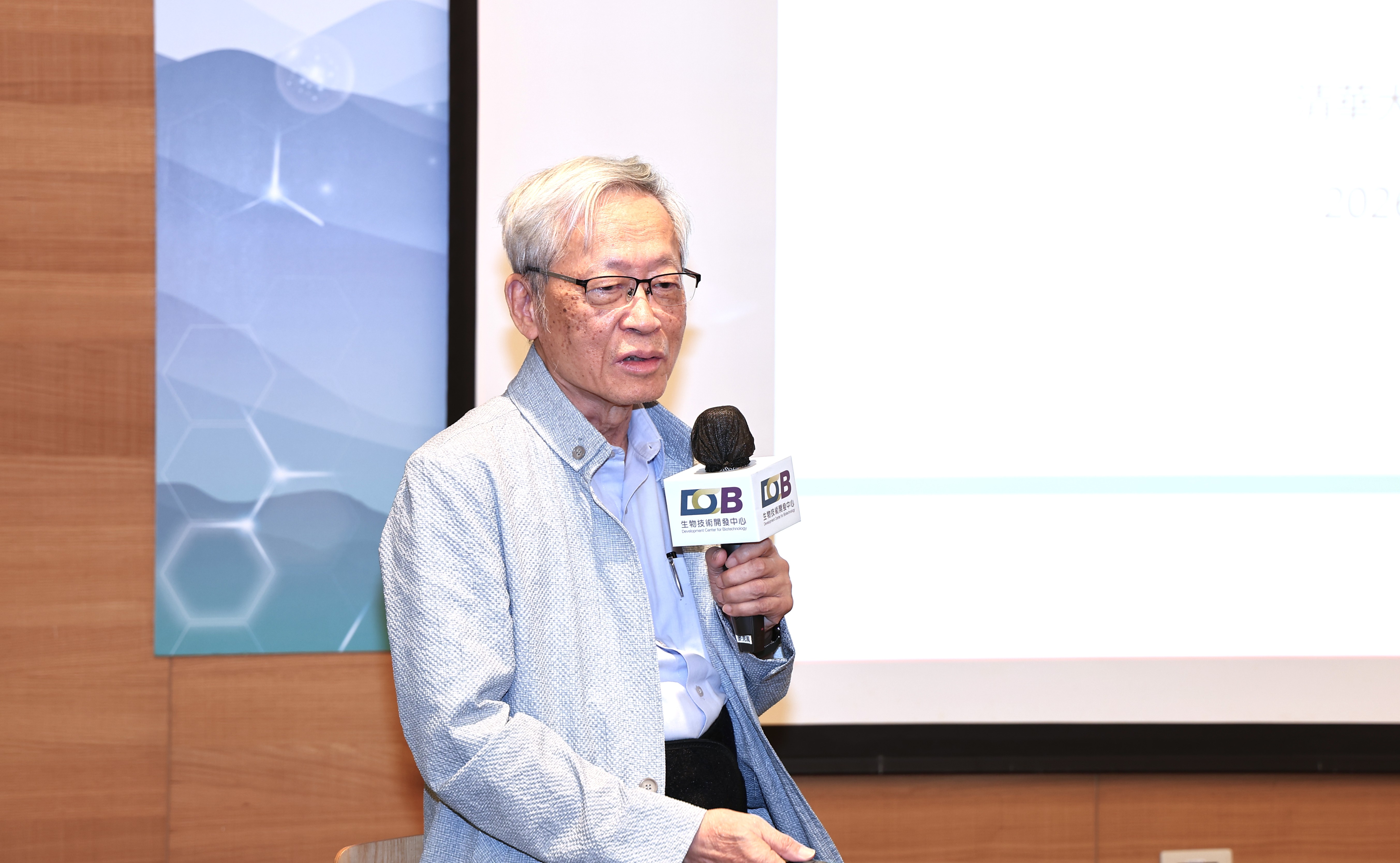 圖片中可見一名戴著眼鏡的年長男子站在室內活動現場發表演講，他穿著淺藍色襯衫與灰藍色背心，胸前掛著工作證，手持黑色麥克風。背景有大型投影螢幕、藍色圖案、木質牆面與明亮燈光，整體呈現正式會議、論壇或學術交流活動的氛圍。